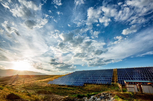 Multiple solar panels in a rural pasture under a partly cloudy blue sky with a rising sun in the background.
